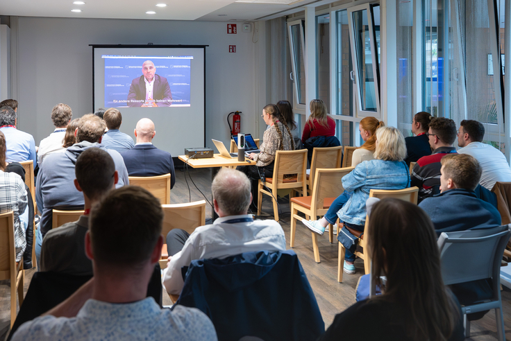 Das Bild zeigt Menschen von hinten, die in Stuhlreihen sitzen. Vor ihnen steht eine Leinwand, auf der ein Video mit einem Grußwort von Niedersachsens CIO Dr. Alexander Georgiadis läuft.