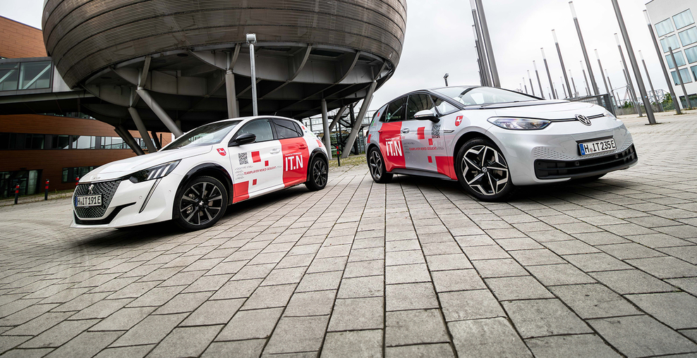 Im Vordergrund parken zwei elektrische PkW mit IT.Niedersachsen-Branding auf gepflastertem Untergrund. Im Hintergrund ist das auf dem Expo-Gelände Hannover befindliche Gebäude 