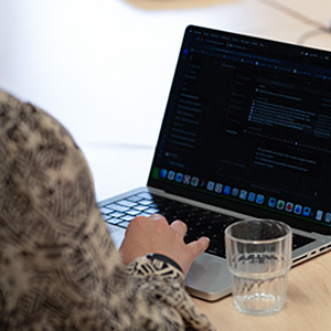 Das Bild zeigt einen aufgeklappten Laptop, daneben ein Glas Wasser und im Vordergrund die Schulter, den Arm und die Hand einer Person, die den Laptop bedient.