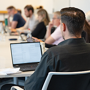 Eine Person von hinten, die an einem Tisch mit einem aufgeklappten Laptop sitzt. Im Hintergrund unscharf weitere Personen, die mit Laptops an Tischen sitzen.