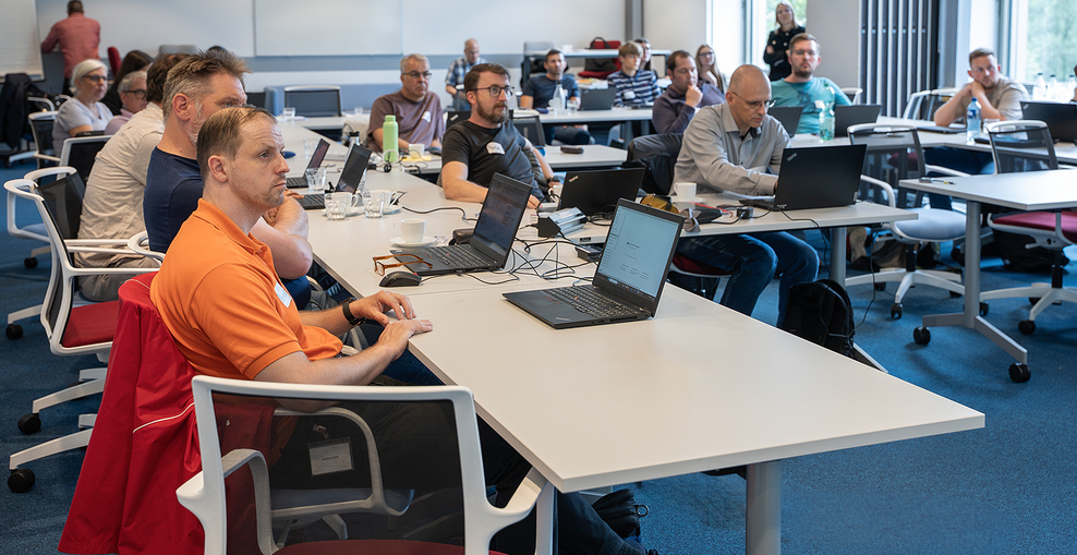 Teilnehmende des Copilot Worklab sitzen mit Laptops an Tischen in einem Konferenzraum.